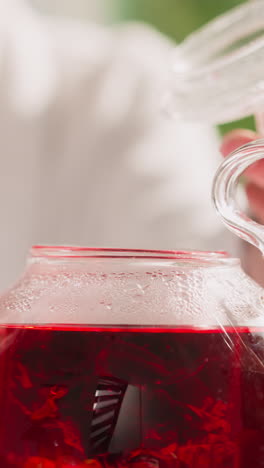una mujer agita té de hibisco en una tetera de primer plano. una mujer prepara una bebida de hierbas rojas para el desayuno en la mesa por la mañana. bebida orgánica saludable casera
