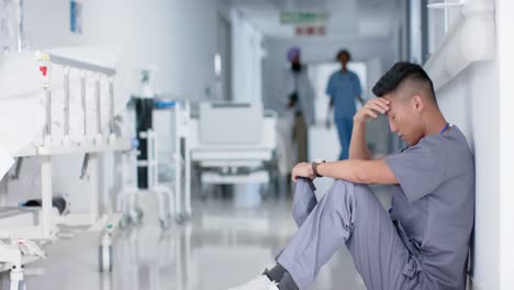 Stressed-asian-male-surgeon-sitting-and-leaning-on-wall-in-hospital-in-slow-motion