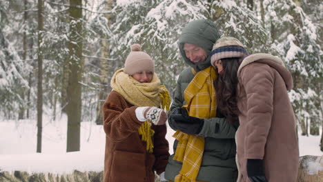 vater, mutter und tochter in winterkleidung, die in einem verschneiten wald auf das smartphone schauen