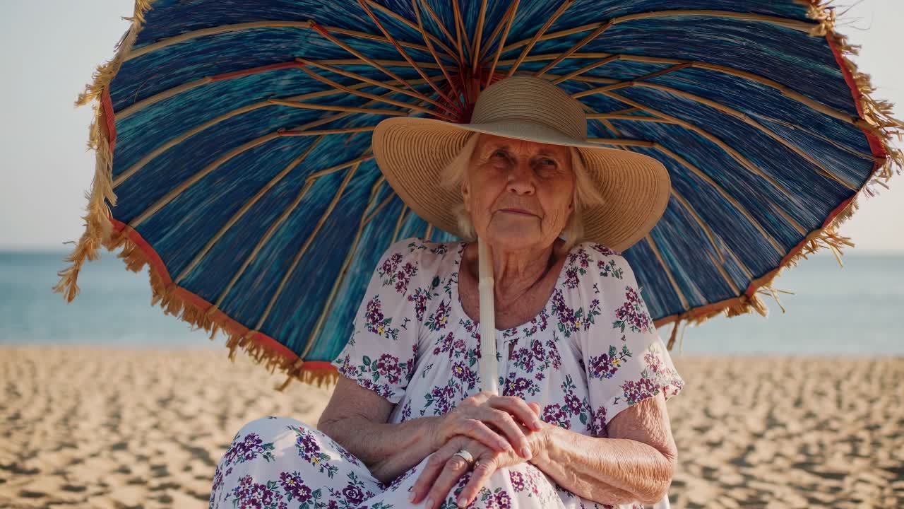 Serene senior woman sitting on sandy beach under blue parasol, enjoying peaceful moment during summer vacation, embracing the tranquility of coastal life