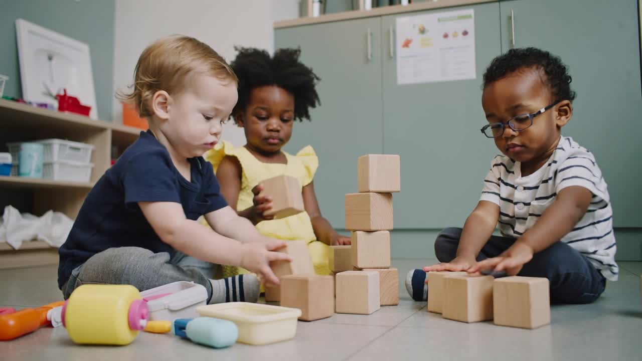 Reaching left child, three toddlers stacking in playroom with blocks, yellow dress practicing motor