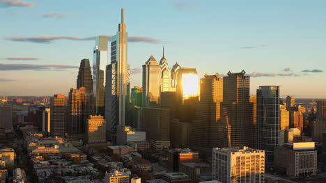 aerial drone pull away view of the downtown philadelphia skyline featuring tall, glass skyscrapers at sunset with golden light and blue summer skies showing the comcast technology center