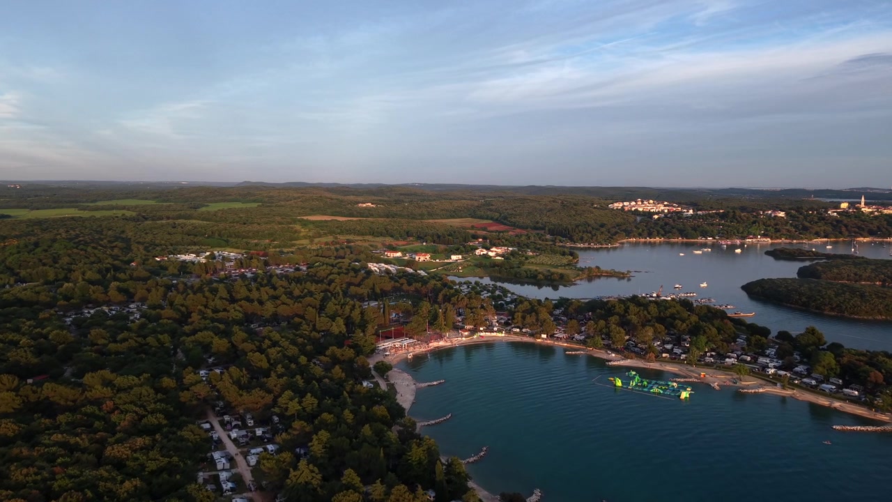 Metsäinen lahti isännöi järvenrantaleirintäaluetta Istriassa, Kroatiassa. Ilmatäytteinen vesipuisto kelluu hiekkarannan lähellä kultaisen ilta-auringon alla, ja taustalla näkyy kaukainen kukkulakylä. Laaja ilmakuva dronella.