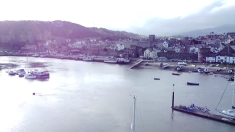 Idyllic-Conwy-castle-and-harbour-fishing-town-coastal-waterfront-aerial-reverse-over-boats-below