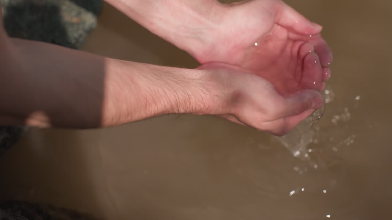 hairy hand scooping muddy river water repeatedly with gentle motion, droplets falling back into brown flowing current, rustic outdoors survival vibe with textured skin details and natural lighting