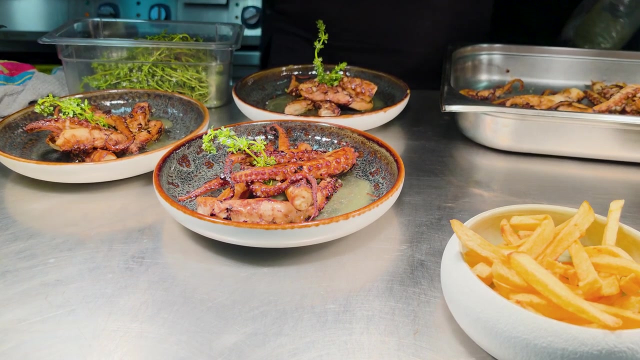 Chef plating grilled octopus tentacles dish in commercial restaurant kitchen squeezing lemon juice on fine dining seafood meal presentation for guests