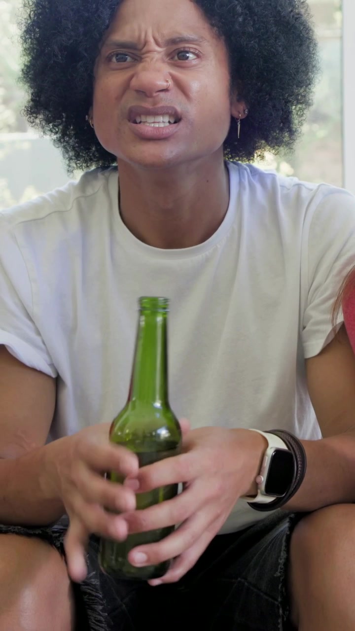 Vertical video: Leaning man in white tee holding green bottle at home, reacting to off-camera event