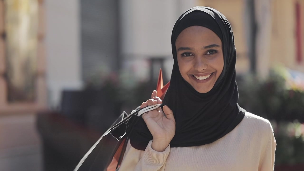 Hymyilevä nuori nainen hijabissa kantaa ostoskasseja ulkona