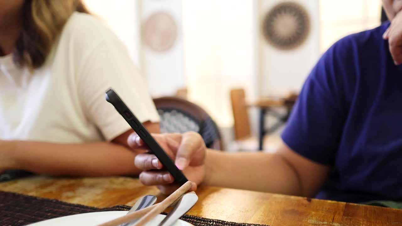 Two individuals sit side by side at a sunlit wooden table, one holding a smartphone, in a casual indoor setting with warm natural lighting