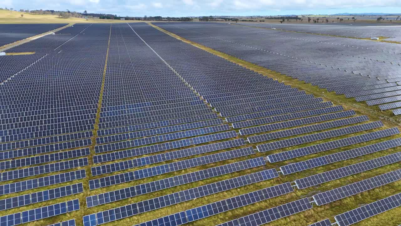 Dronekamera svæver over et stort solcelleanlæg i et landligt landskab i dagslys, der viser infrastruktur for vedvarende energi og geometriske panelmønstre.