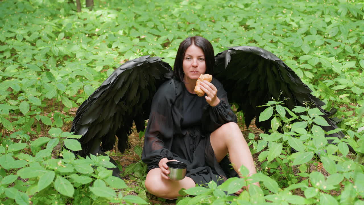 Una donna misteriosa vestita da fata oscura con ali nere piumate siede sul suolo di una foresta circondata da piante verdeggianti, mangiando un dolce e tenendo in mano una tazza di metallo, fondendo eleganza mistica
