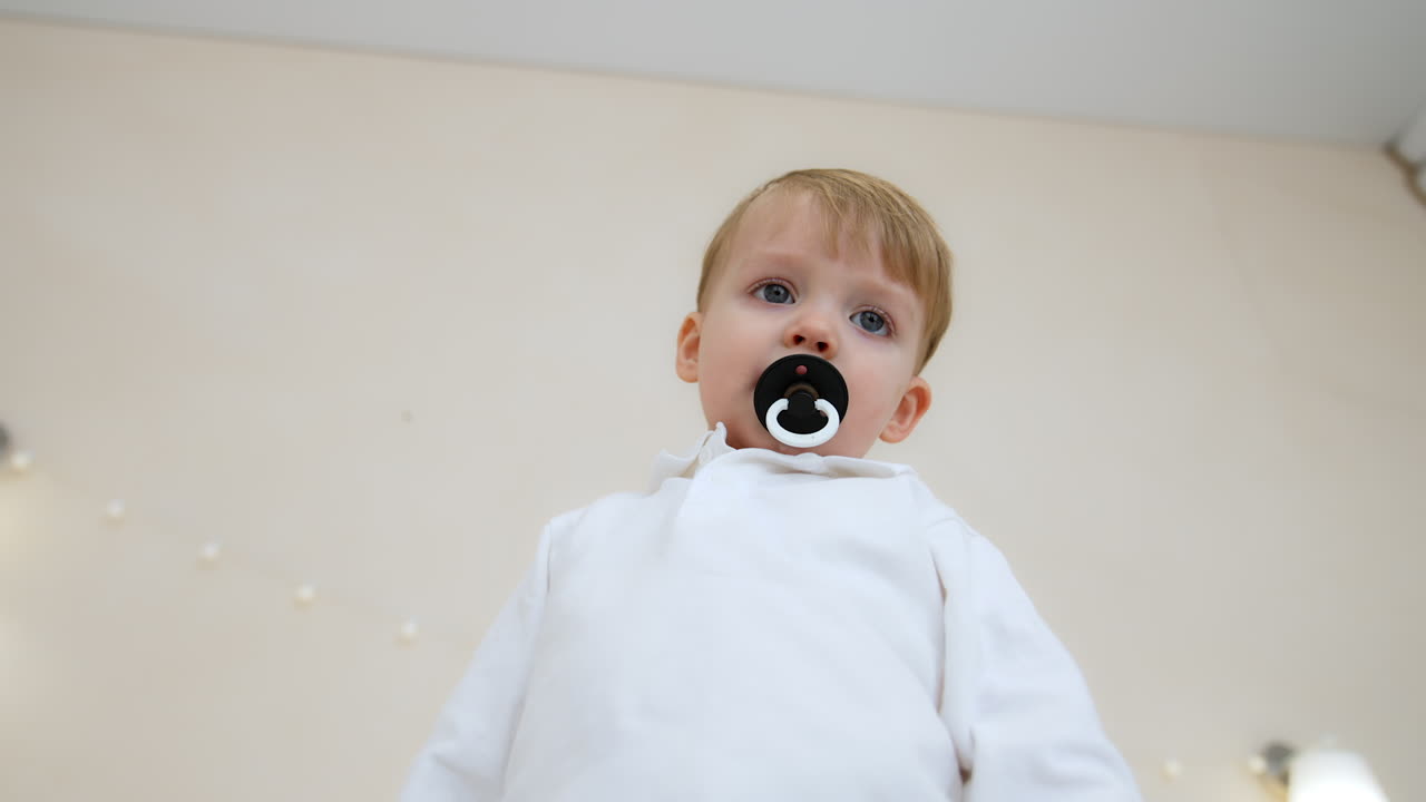 Toddler boy in white shirt sucking a pacifier. Low angle view at the calm Caucasian baby.
