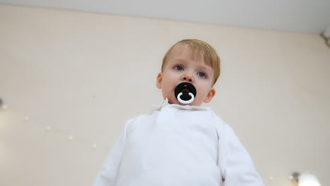 toddler boy in white shirt sucking a pacifier. low angle view at the calm caucasian baby.