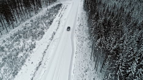 aerial tracking shot of car driving on snowy road in forest landscape during winter day