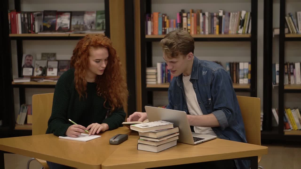 Young male and female students talking, explaining course work study together in library. Actively discussing something sitting by wooden table with a book. Front view
