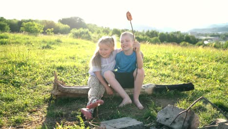 Sister hugging brother while they have conversation sitting on log and roasting a  sausages on the sticks over the campfire. Happy family or outdoor picnic activities concept image.