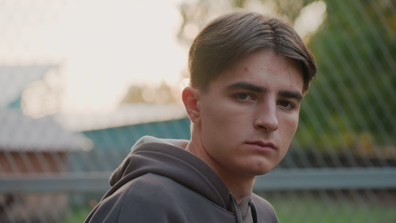 Adolescente presso una recinzione metallica al tramonto, studente con felpa con cappuccio guarda pensieroso in lontananza; il primo piano rivela un'espressione contemplativa, fienile di periferia sullo sfondo, retroilluminazione soffusa e toni cinematografici caldi.