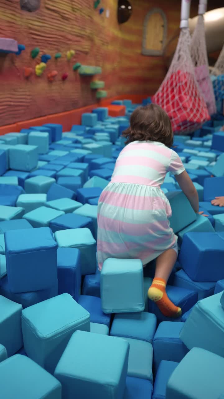 Little girl in a striped dress has fun climbing and crawling through a large pit filled with soft blue foam blocks at an indoor playground and activity center