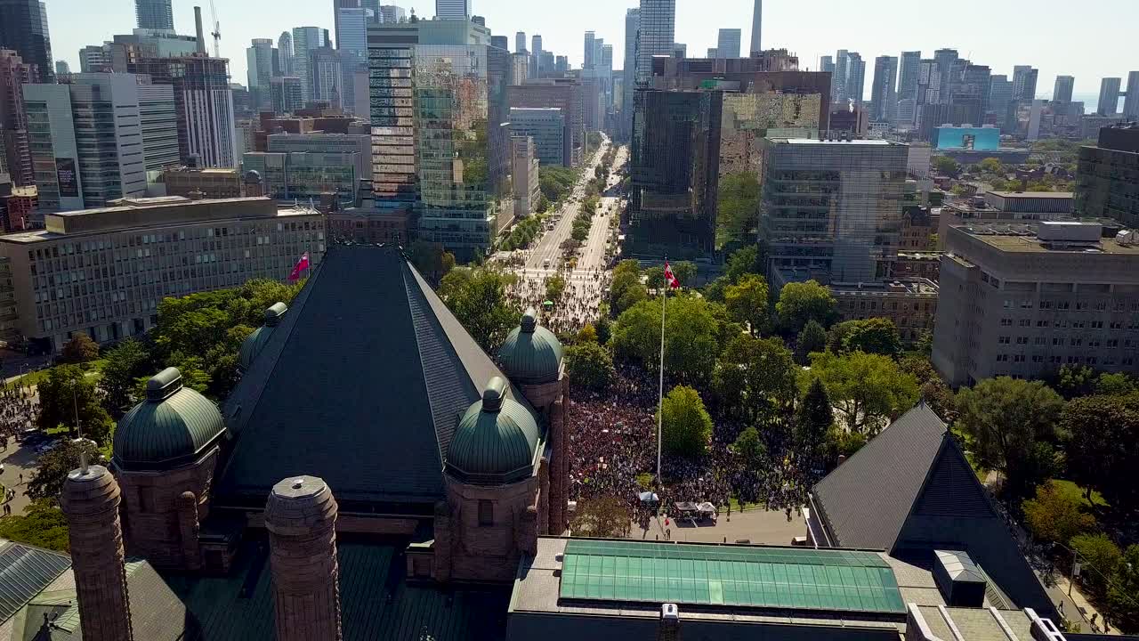 Aerial view of downtown Toronto and Ontario Legislature, wide daytime fixed shot in 4k. Old building looms over crowd of people on front lawn as city skyscraper buildings tower in panorama background