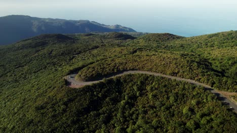 Drohnenaufnahmen-Einer-Kurvigen-Straße-In-Grünen-Bergen,-Fahrendes-Auto,-Meerblick-Im-Hintergrund,-Luftaufnahmen-Von-Madeira