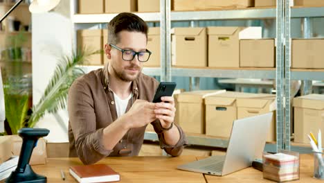 caucasian designer man in eyeglasses sitting at desk texting on mobile phone in clothing shop warehouse