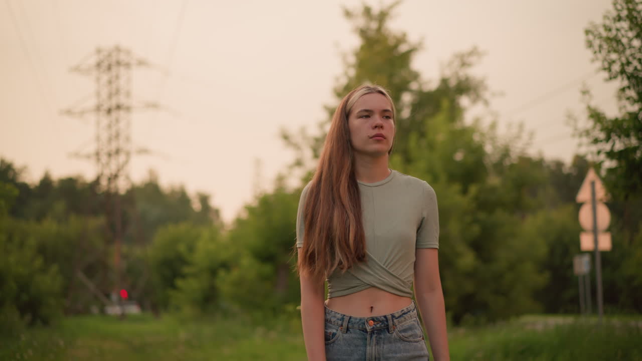 Young woman with long hair walking along grassy path, looking tired and contemplative. Power lines and blurred background with passing car