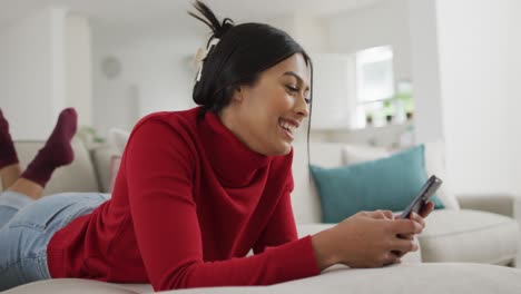 Video-of-happy-biracial-woman-in-hijab-lying-on-sofa-and-using-smartphone