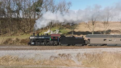 una vista lateral aérea en cámara lenta, de un tren de pasajeros de vapor que se acerca, soplando humo, mientras viaja por el campo, en un día soleado de invierno, en un día soleado de invierno