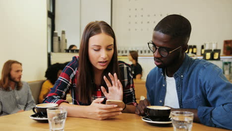 caucasian woman and african american man watching a video on smartphone in a cafe