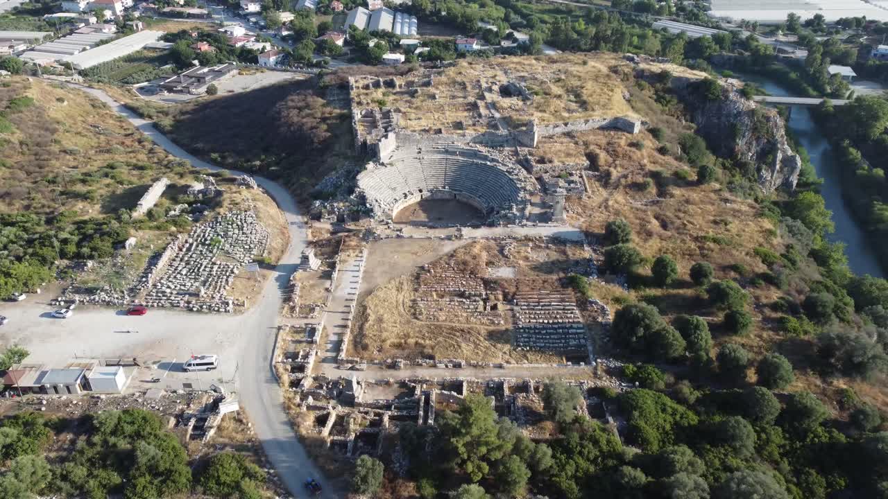 Aerial drone view of restored amphitheater and grid like stone ruins beside winding river at ancient Patara in Kas Antalya Province under bright daylight across dry terrain with scattered greenery