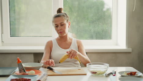 food preparer carefully observes banana before cutting on wooden board in bright kitchen, ensuring it is neatly washed, with kitchen tools, bowls, and ingredients arranged around clean workspace