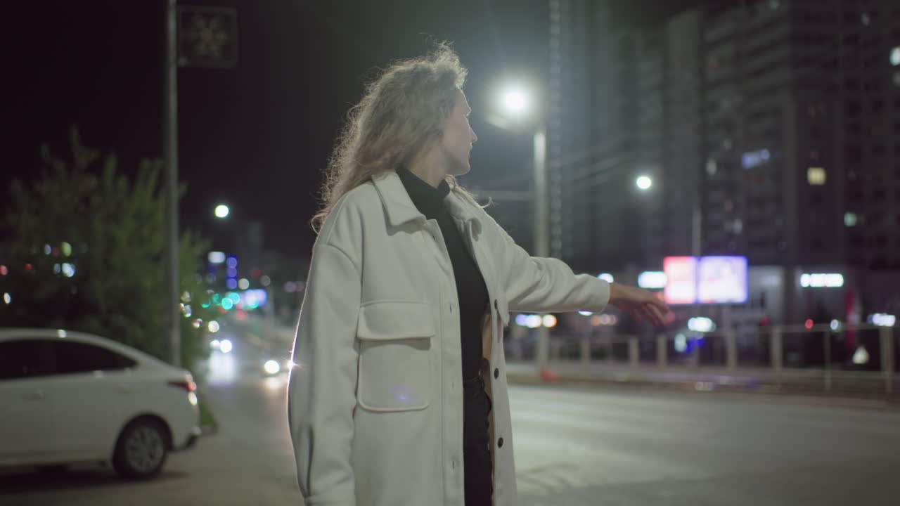 Female marketer closes late from work, stands by roadside stopping car under streetlights as another car drives past in urban environment with parked white car in background and blurred city lights