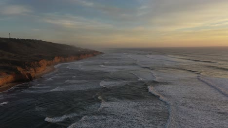 una impresionante toma de drones volando sobre los brumosos acantilados con las olas rompiendo debajo, capturando el poder puro de la naturaleza y la calidez del sol poniente