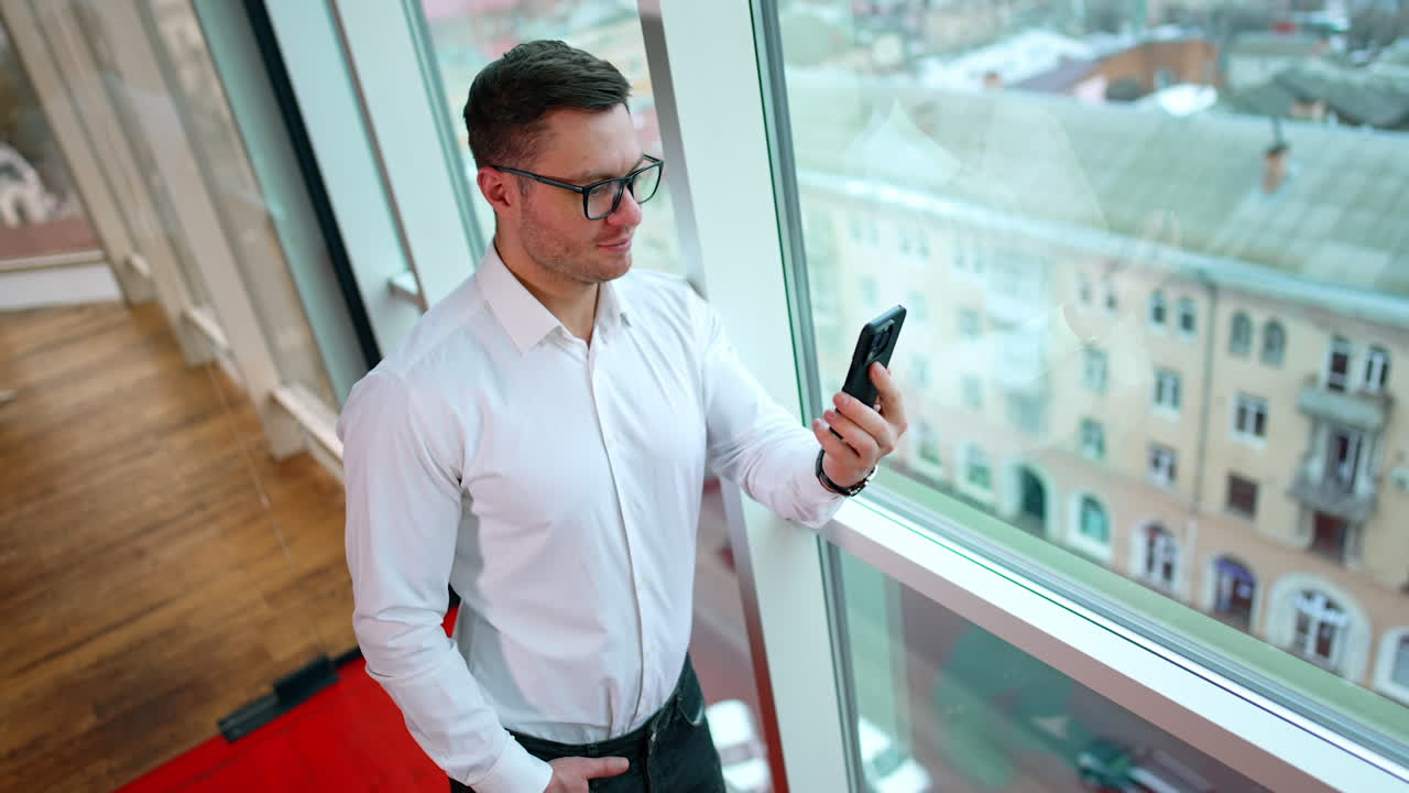 Confident male entrepreneur talking to partners via video chat. Man in glasses and white shirt using smartphone in work. Top view.