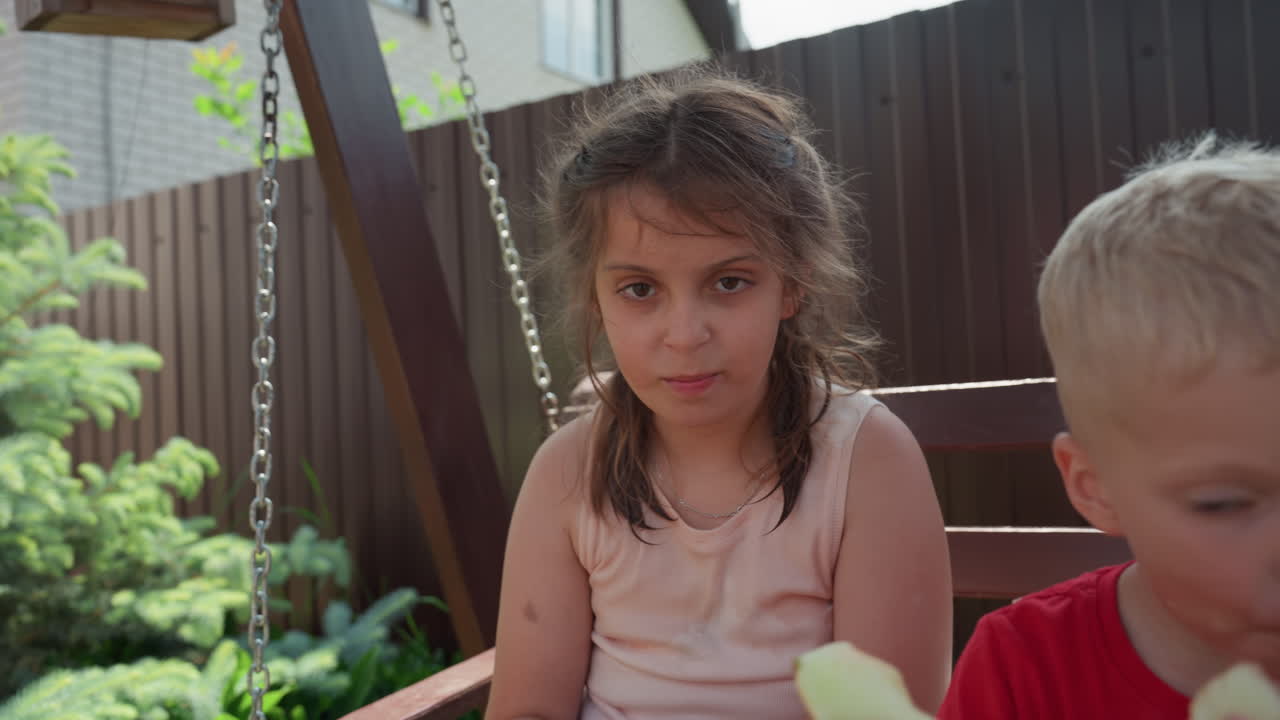 Child Enjoying Summer, Girl On Swing Tasting Fruit Outdoors, Child On Swing Savoring Melon With Sibling In Garden, Juvenile Girl Swinging And Tasting Melon Slices Amid Lush Green Environment