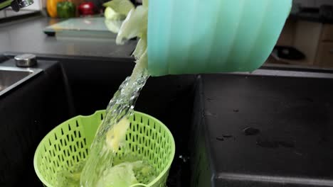 pouring lettuce leaves with water into plastic strainer in the kitchen sink