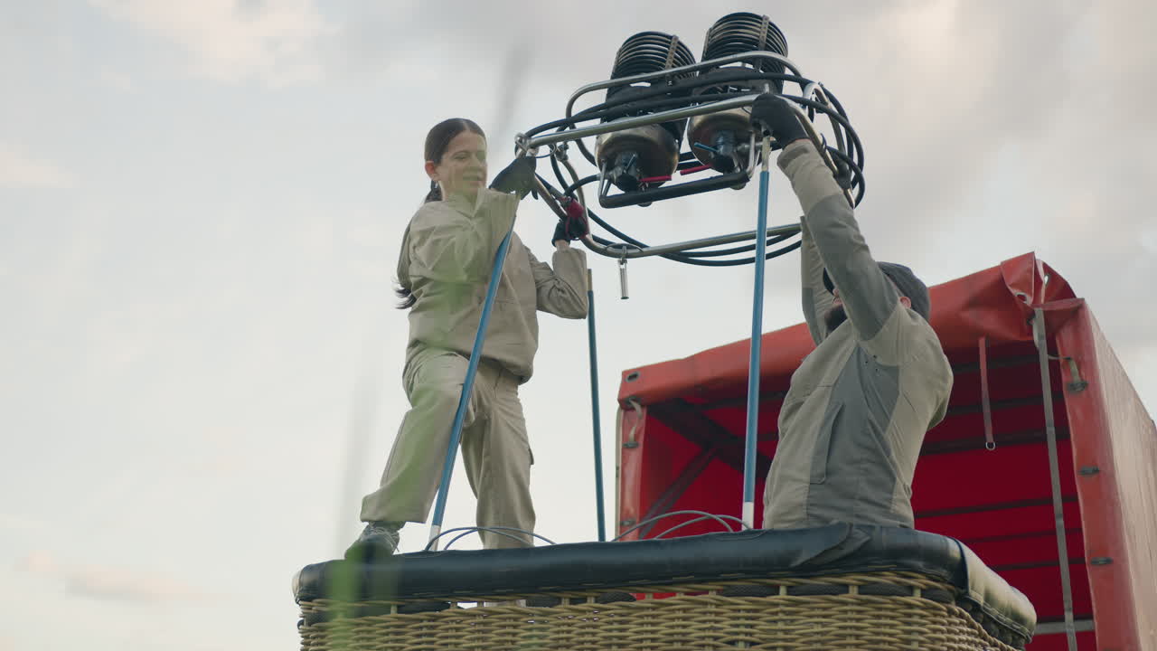 Man seated inside wicker basket holds metal burner while woman standing above helps mount unit onto upright poles during hot air balloon assembly under soft daylight with red truck in background