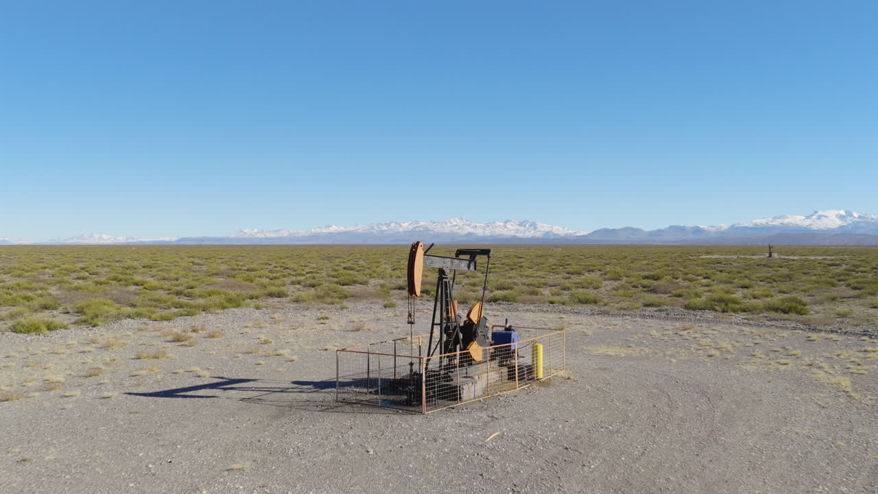 Aerial shot of an oil pump jack in Vaca Muerta transitioning to a vast Patagonian desert and the Andes mountains in the distance.