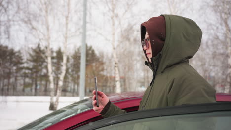 boy in green winter jacket and sunglasses taking selfie with smartphone while standing beside red car during light snowfall, trees and blurred moving cars in background on cold snowy day
