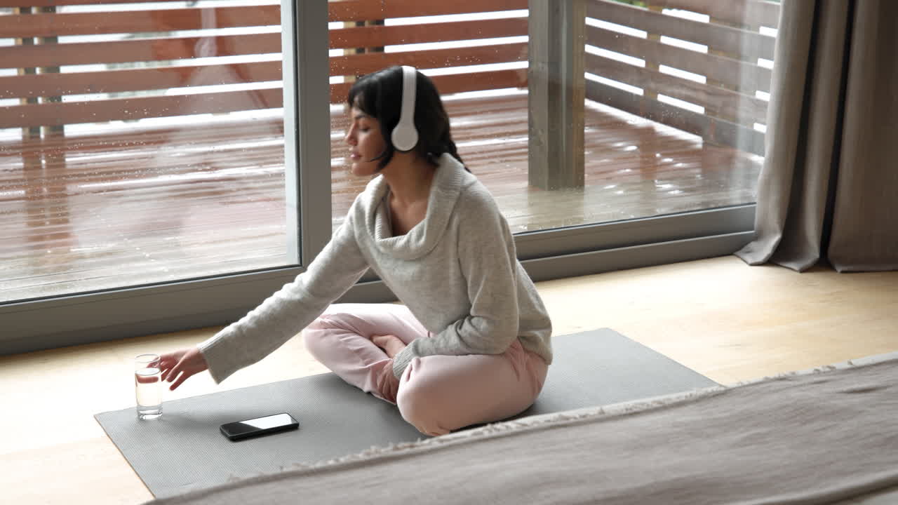 Relaxing at home, woman drinking water with headphones on yoga mat