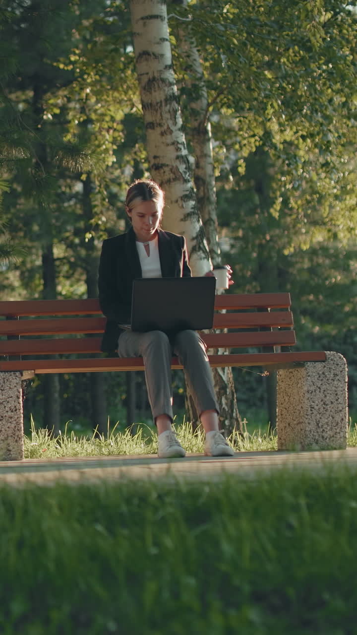 Freelance seduto all'aperto su una panchina di legno mentre lavora sul sistema e sorseggia un succo, circondato dal verde lussureggiante di un parco con la calda luce del mattino