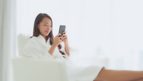 Relaxing-in-a-bathrobe-while-sitting-in-a-comfortable-chair-in-a-hotel-suite,-a-pretty-young-woman-focuses-on-her-smartphone-and-texting