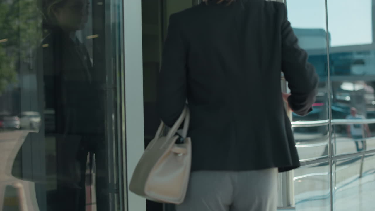 Office worker walks into office building dressed professionally with glass door reflecting urban surroundings, parked cars, and street, focus on back view modern corporate environment