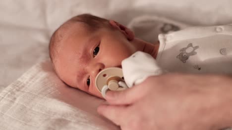 male calming upset newborn baby boy laying on the sofa by putting dummy in his mouth