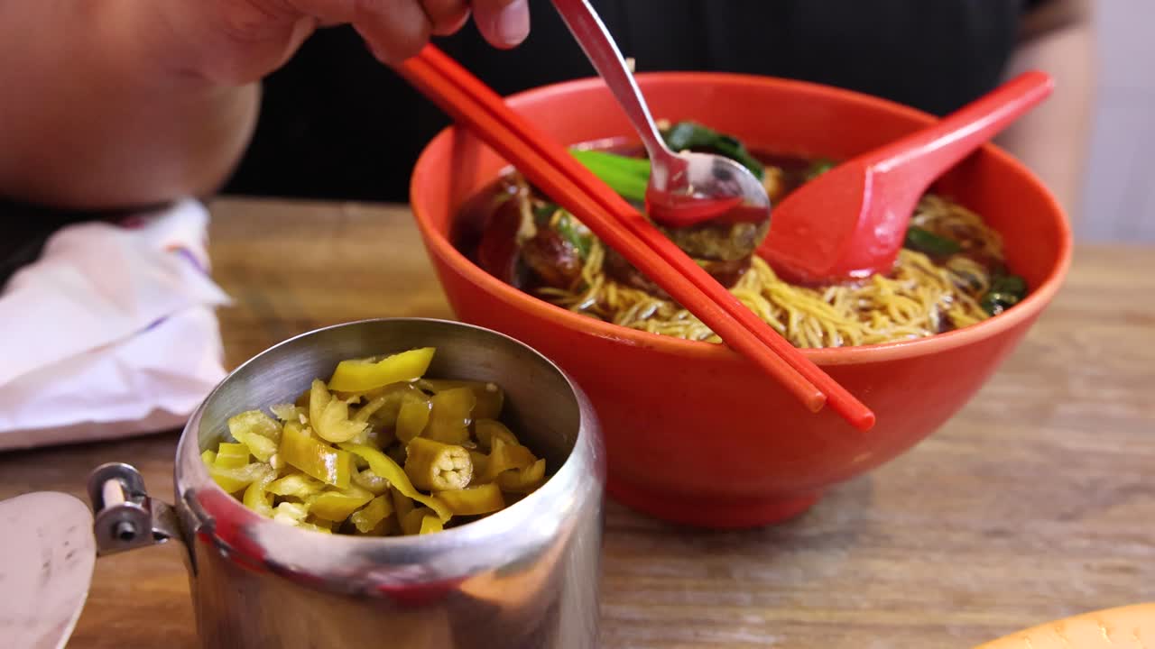 A hand uses a spoon to scoop pickled chili from a metal container and adds it to a bowl of noodles in a casual dining setting with warm lighting