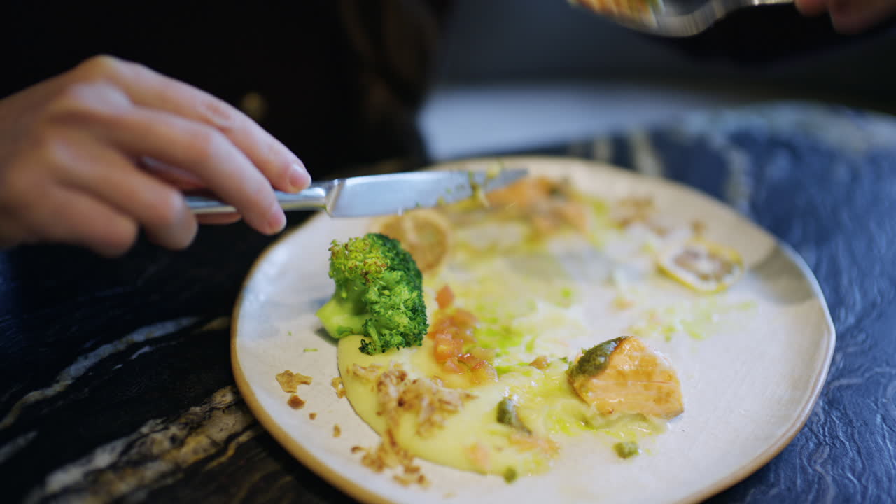 Primo piano di mani che usano coltello e forchetta per tagliare un filetto di salmone servito con broccoli e salsa.