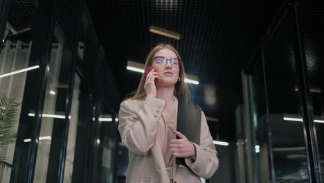 Smiling-businesswoman-looking-on-cellphone-indoors.-Surprised-business-woman-reading-message-on-mobile-phone-in-office-corridor.-woman-looking-smartphone-screen-in-business-center.-walking-office