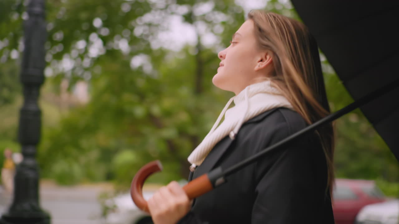 Gorgeous lady holding umbrella running softly as hair blows wildly across face wearing black coat and white hoodie near parked cars with blurred green trees in background