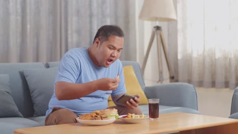 a fat asian man having french fries with ketchup and playing game on smartphone while eating fast food on a sofa in the living room at home