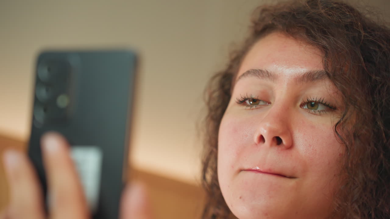 Hotel room woman applying lip gloss using phone mirror warm morning light curly hair freckles closeup focused expression traveler prepping for meeting casual chic grooming routine glossy pink finish